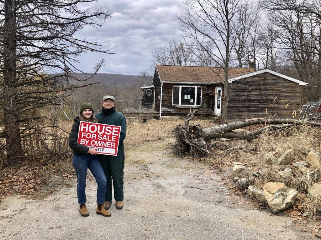 Bea and her father stand outside her newly purchased home, holding a red "for sale" sign.