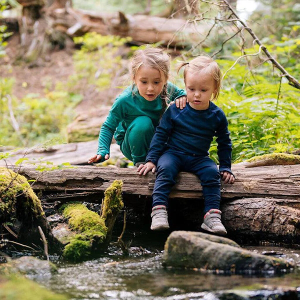 Two young blond children wear matching wool long-sleeved outfits, one in navy and one in teal, and climb a log over a small spring.