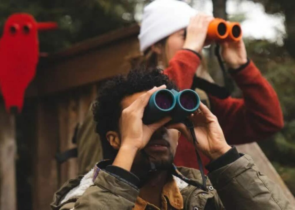 Two people stand outside, dressed for cold weather, and look through binoculars. 
